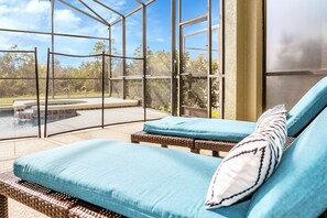 Área de estar ao ar livre ensolarada, com móveis de vime e vista para uma piscina e vegetação.