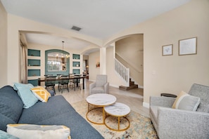 A spacious living area with a grey sofa, a grey armchair, and a small round table with marble tops. There is a staircase leading to an upper floor, and a dining area with a table and chairs is visible in the background. - Emerald Island 8581 (Kissimmee)