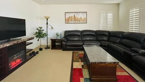 Ein Wohnzimmer mit schwarzem Ledersofa, Flachbildfernseher, Couchtisch, Stehlampe, Blumentopf und Stadtbild an der Wand.