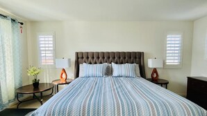Ein Schlafzimmer mit einem großen Bett, zwei Nachttischen, einem Beistelltisch, einer Topfpflanze und einem Fenster mit Jalousien.