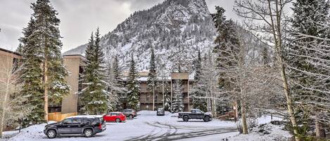 Un estacionamiento nevado con vehículos, una montaña al fondo y un edificio de varios pisos.