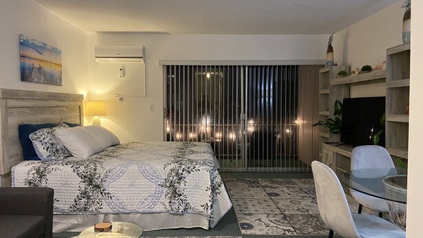 Quarto de hotel com cama, mesa de centro de vidro, cadeira, televisão e varanda com luzes.