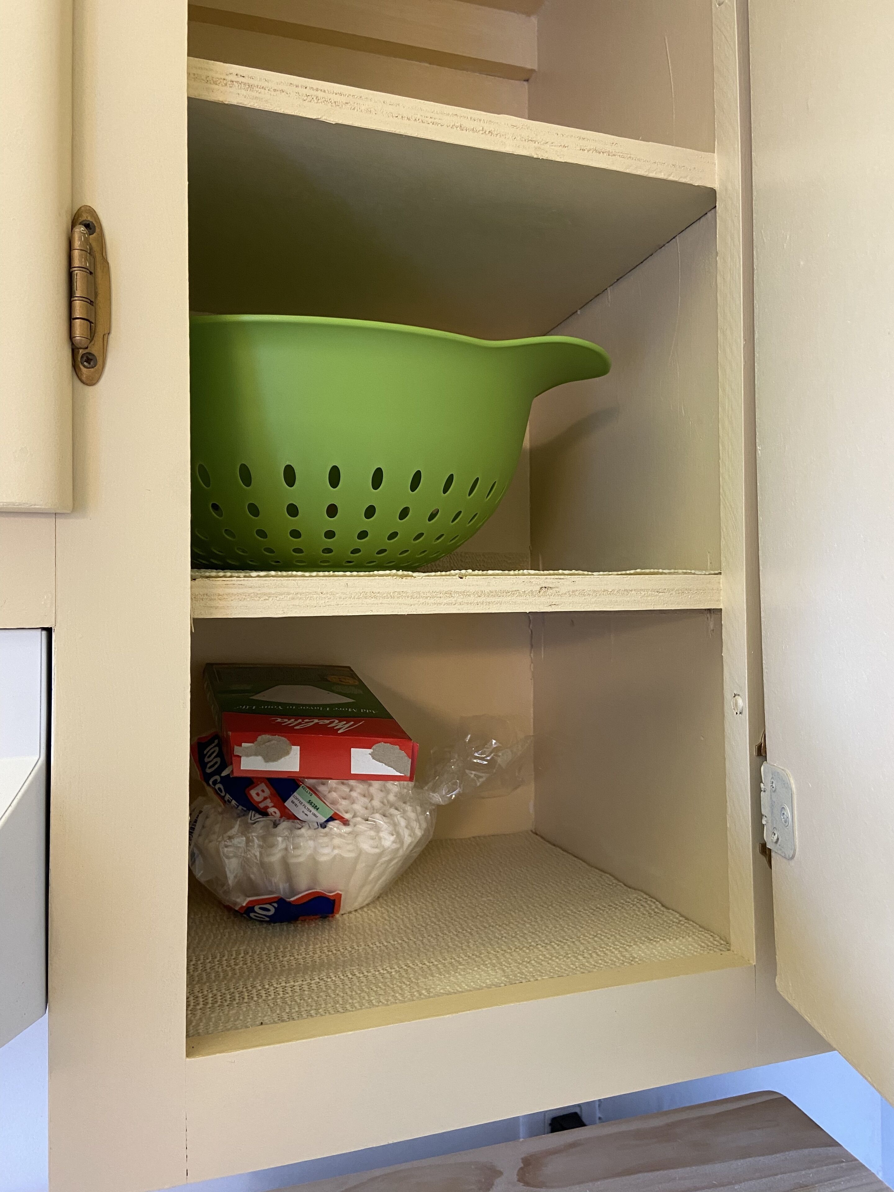 A kitchen cabinet with a green colander on the top shelf and a bag of white noodles on the bottom shelf.