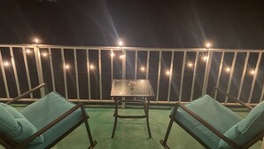 Uma varanda com duas cadeiras azuis acolchoadas e uma mesa pequena, cercada por um corrimão branco e luzes de fada.