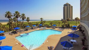 A resort pool area with lounge chairs and umbrellas, overlooking a beach and a tall building. - Heated Pool + Gulf View: Galvestonian Resort Condo (Galveston)