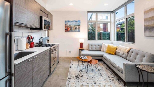 A modern kitchen with a built-in stove, microwave, and stainless steel appliances. There is a grey sofa with yellow cushions and a small wooden side table. A large window provides a view of the outside. - Walkable in Heart of Ballard (Seattle)