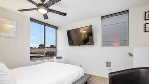 A modern bedroom with a bed, a ceiling fan, a TV, and a window with a view of buildings. - Walkable in Heart of Ballard (Seattle)