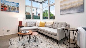 A modern living room with a grey sectional sofa, a small round coffee table, a side table, and a large window offering a view of the outside. - Walkable in Heart of Ballard (Seattle)