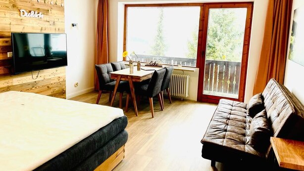 Una habitación con una cama, un televisor fijado a una pared de madera, una mesa de comedor con sillas, un sofá y un balcón con vistas a árboles.