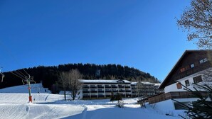 Estación de esquí nevada con un edificio de varios pisos, telesilla y montañas al fondo.