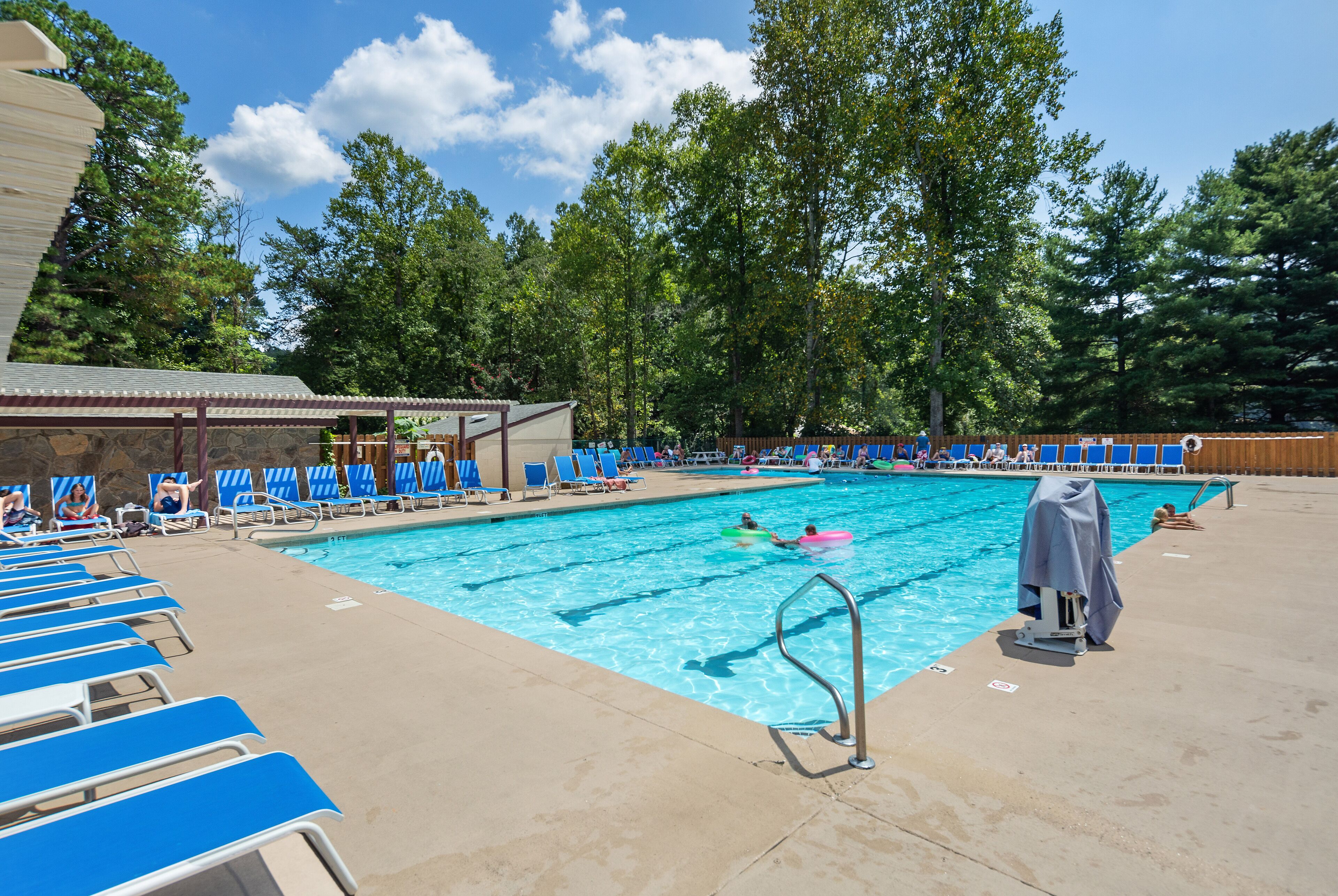 A swimming pool with blue lounge chairs, a covered area, and trees in the background.
