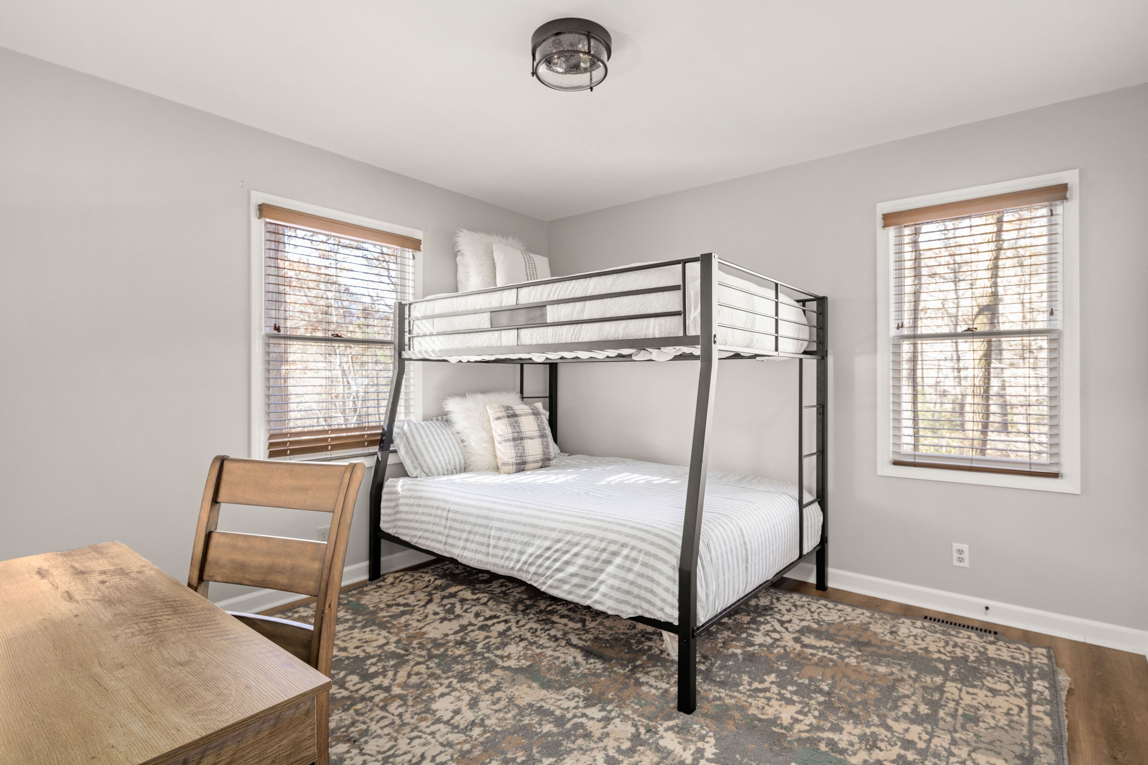 A bunk bed room with a wooden chair, a patterned rug, and two windows with blinds.