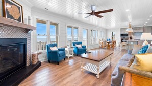 A spacious living room with a fireplace, blue armchairs, a wooden coffee table, and a view of the kitchen and outdoor balcony. - "Porpoise Point" - Oceanfront Home, 5 Bedrooms / 5 Bathrooms, w/ PRIVATE POOL (Holden Beach)