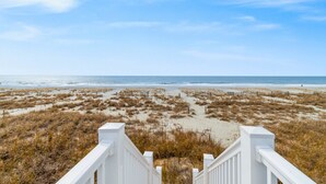 A beach view from a white staircase leading to a sandy area with grass and a wooden fence. - "Porpoise Point" - Oceanfront Home, 5 Bedrooms / 5 Bathrooms, w/ PRIVATE POOL (Holden Beach)