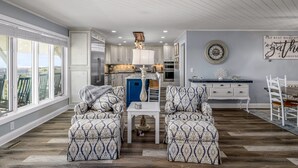 A spacious living area with a large window, a dining table with chairs, a blue cabinet, and a decorative wall clock. - "All Hands On Deck" Oceanfront with elevator 5 bedrooms, 5 bathrooms. (Holden Beach)