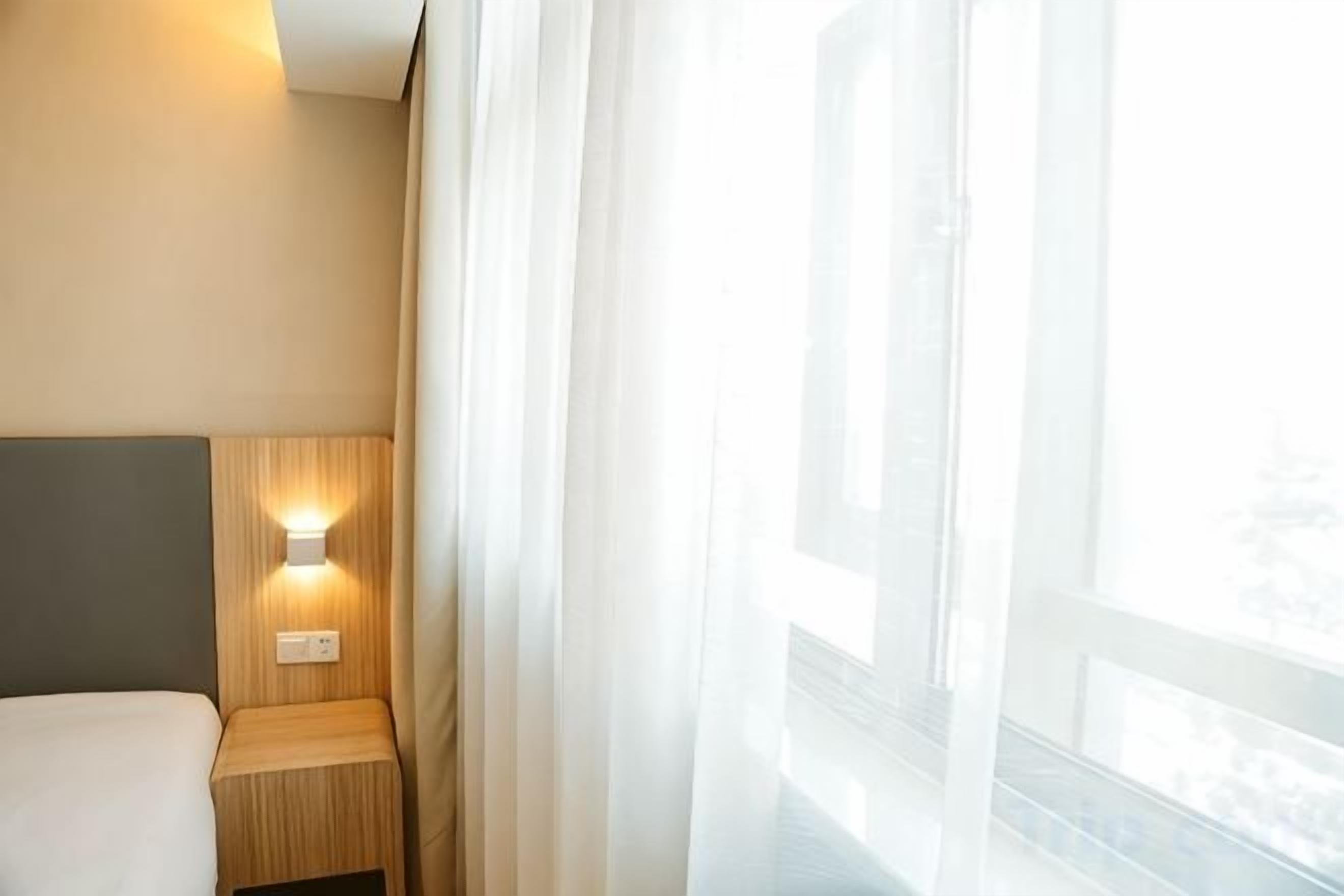 A hotel room with a bed, a nightstand with a lamp, and a window with sheer curtains.