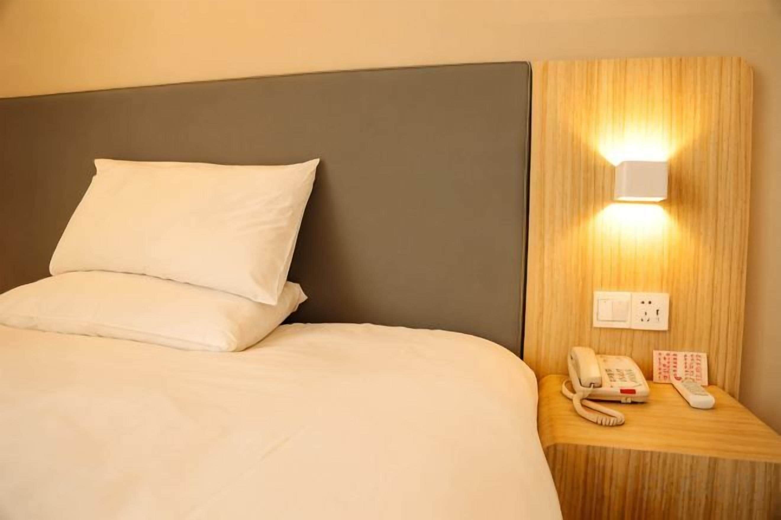 A hotel room with a bed, a bedside table with a phone and a remote control, and a wall-mounted light fixture.