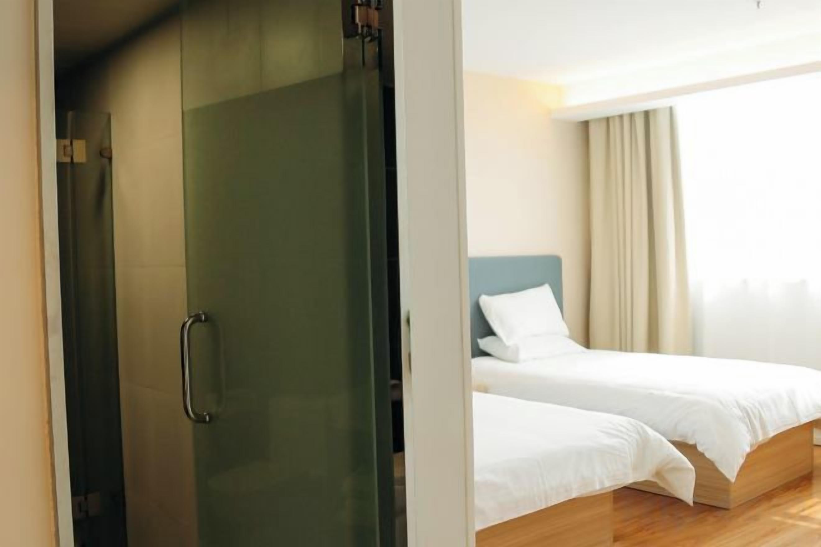 A hotel room with a glass door leading to a shower area, a bed with white bedding, and a view of the outdoors.