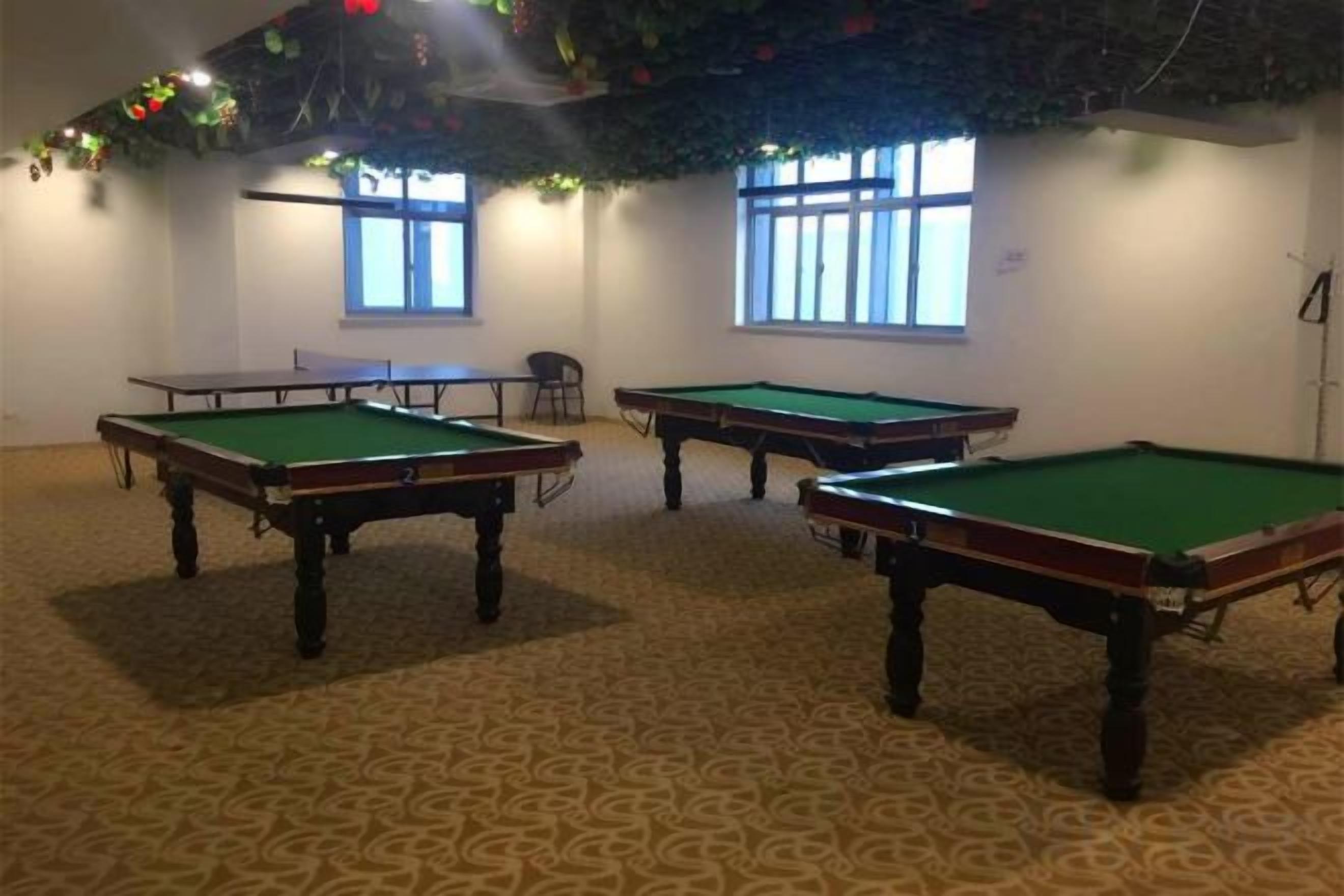 A room with three pool tables and a ping pong table, carpeted floor, and windows with blinds.