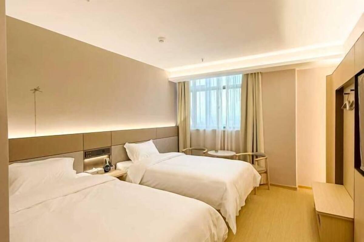 A hotel room with two beds, a bedside table with a lamp, a small round table, and a window with curtains.