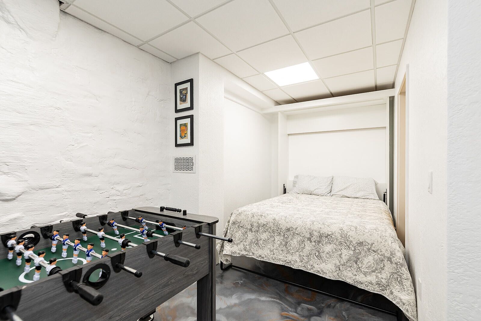 Une pièce avec une table de baby-foot, un lit recouvert d’une couvre-lit à motifs et un mur blanc à la texture particulière.
