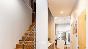Un interior moderno con una escalera de madera, una zona de cocina y un pasillo que conduce a una puerta.