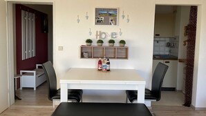 A modern living room with a white dining table, black chairs, and a bench. There is a kitchen visible through an open doorway.