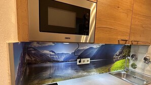 A kitchen with wooden cabinets, a stainless steel microwave, a sink, and a backsplash with a scenic landscape mural.