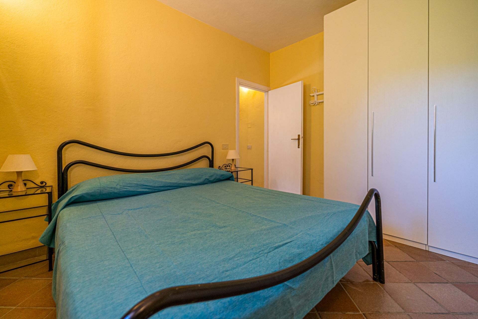 Uma cama de solteiro com estrutura de metal e colcha azul num quarto com paredes amarelas e porta branca.