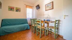 Een woonkamer met een smaragdgroene bank, een houten eettafel met vier stoelen, een aan de muur bevestigde televisie en een deur met een greep.