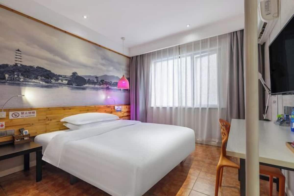A hotel room with a bed, a desk, a chair, a window with curtains, a wall-mounted TV, and a wall with a scenic painting.
