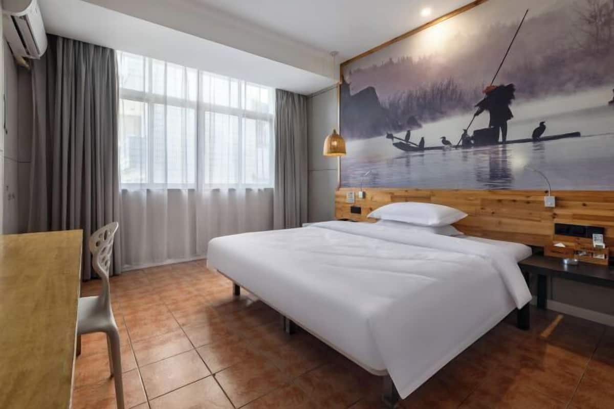 A hotel room with a large bed, a desk, a chair, a painting of a lake scene, and a window with curtains.