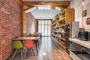 Un pasillo estrecho con pared de ladrillo, vigas de madera en el techo y una zona de comedor con sillas de colores.
