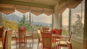 A dining area with wooden chairs and a table, large windows with curtains, and a view of mountains and trees. - Hotel Fotini (Kalamata)
