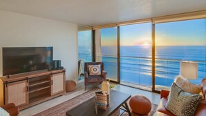 A modern living room with a large TV, a comfortable sofa, a chair, and a view of the ocean through large windows. - WOW 10th Fl Corner Unit * Ocean Views * Pool + Hot Tub (San Diego)