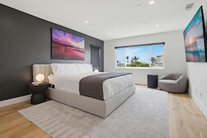 Un dormitorio moderno con una cama grande, una mesita de noche con lámpara, una ventana con vistas y una silla gris.