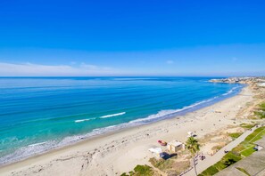 Una playa costera con aguas azules cristalinas, orilla arenosa y algunos edificios al fondo.