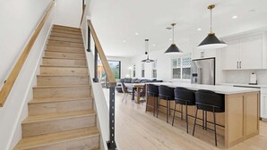 Une cuisine moderne avec un plancher en bois, un îlot central doté de tabourets de bar, un éclairage suspendu et des meubles de cuisine blancs.