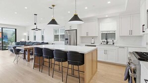 Une cuisine moderne avec des meubles blancs, un îlot central doté de tabourets, un éclairage suspendu et une grande fenêtre.