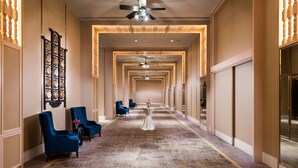 Un couloir d’hôtel avec un tapis à motifs, des fauteuils bleus et une table sur laquelle est posé un vase.