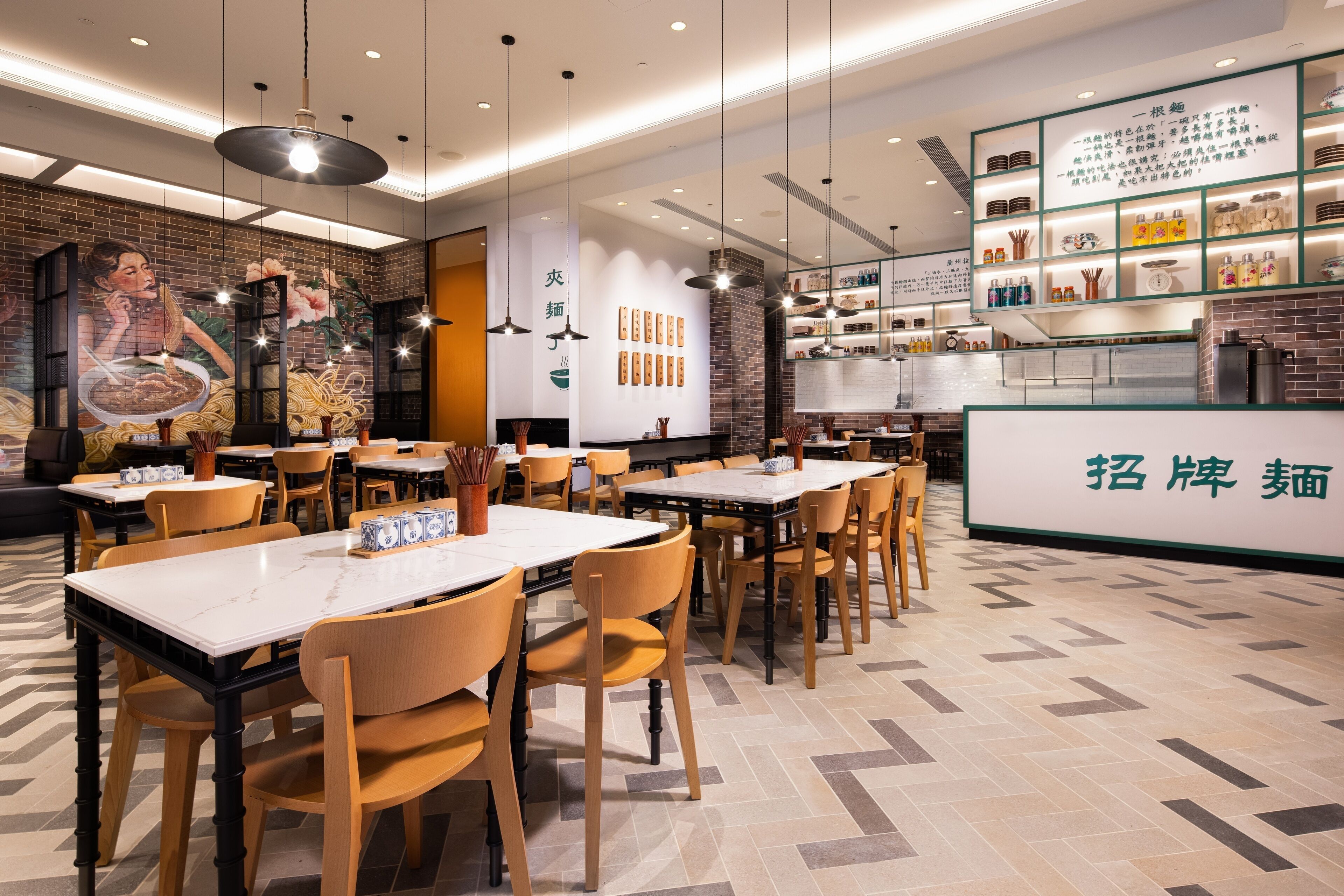 A restaurant interior with wooden tables and chairs, hanging pendant lights, a mural on the wall, and a display of dishes and utensils.