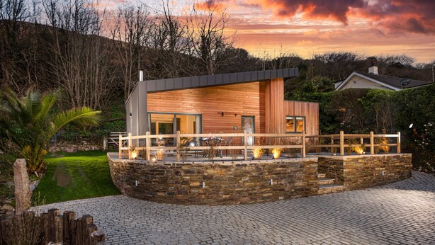 A modern house with a stone wall and wooden deck, surrounded by greenery and a cobblestone driveway.