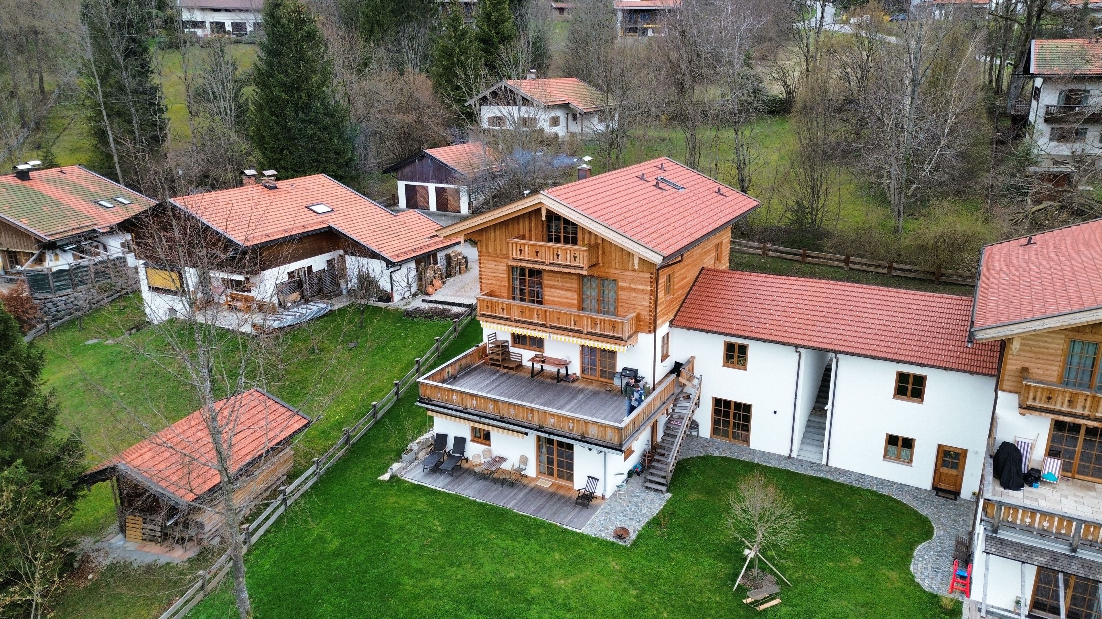 A multi-story wooden chalet with a balcony, surrounded by a well-maintained lawn and neighboring houses.