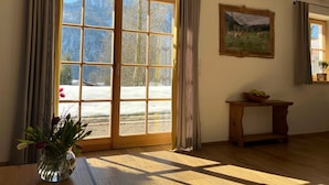 Une pièce dotée d’une grande porte-fenêtre coulissante, d’un banc en bois, d’une petite table sur laquelle se trouve un saladier rempli de fruits, et d’un tableau représentant un paysage, accroché au mur.
