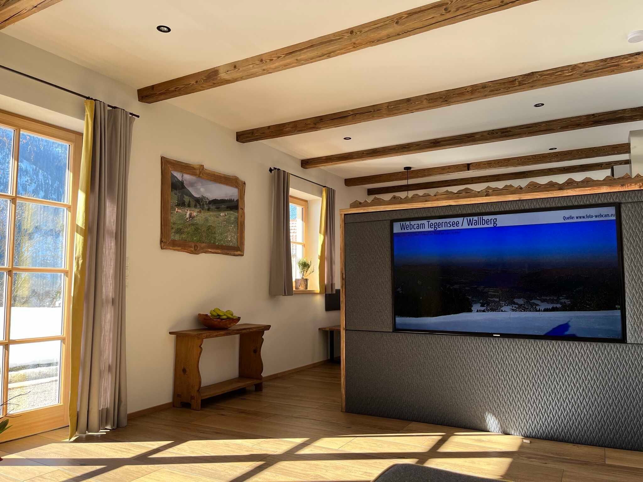 Ein Zimmer mit Holzbalken, einem großen Fenster, einem gerahmten Bild, einem Holztischelement und einem Fernsehbildschirm, der eine Landschaft zeigt.