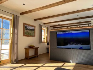 A room with wooden beams, a large window, a framed picture, a wooden console table, and a television screen displaying a landscape.