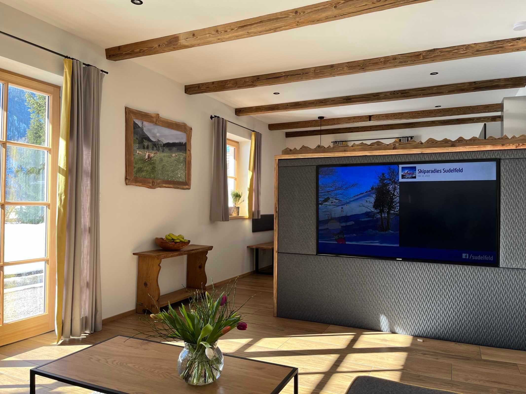 A living room with wooden beams, a TV mounted on a textured wall, a wooden table, and a vase with plants.