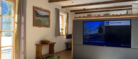 A living room with wooden beams, a TV mounted on a textured wall, a wooden table, and a vase with plants.