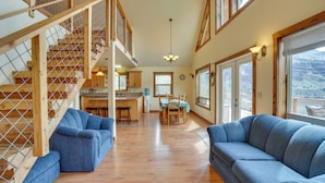 A spacious living area with a wooden staircase, a dining area, and large windows offering a view of the outdoors.