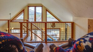 A high-ceilinged room with a sloped roof, a wooden staircase, and a view through large windows.